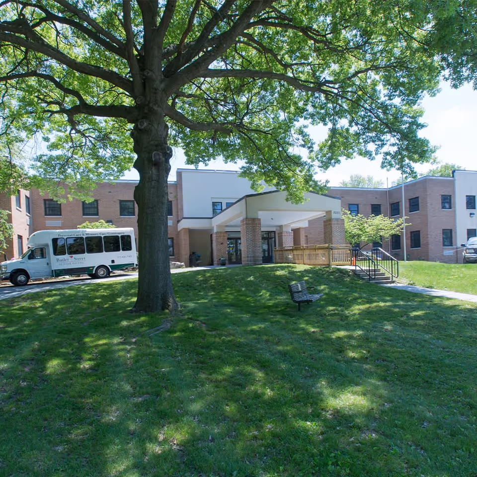 Exterior view of Butler Street Senior Living facility showing a large tree in the foreground, a grassy area with a bench, and the building entrance with a covered drop-off area. A white shuttle bus with Butler Street Senior Living branding is parked near the entrance.