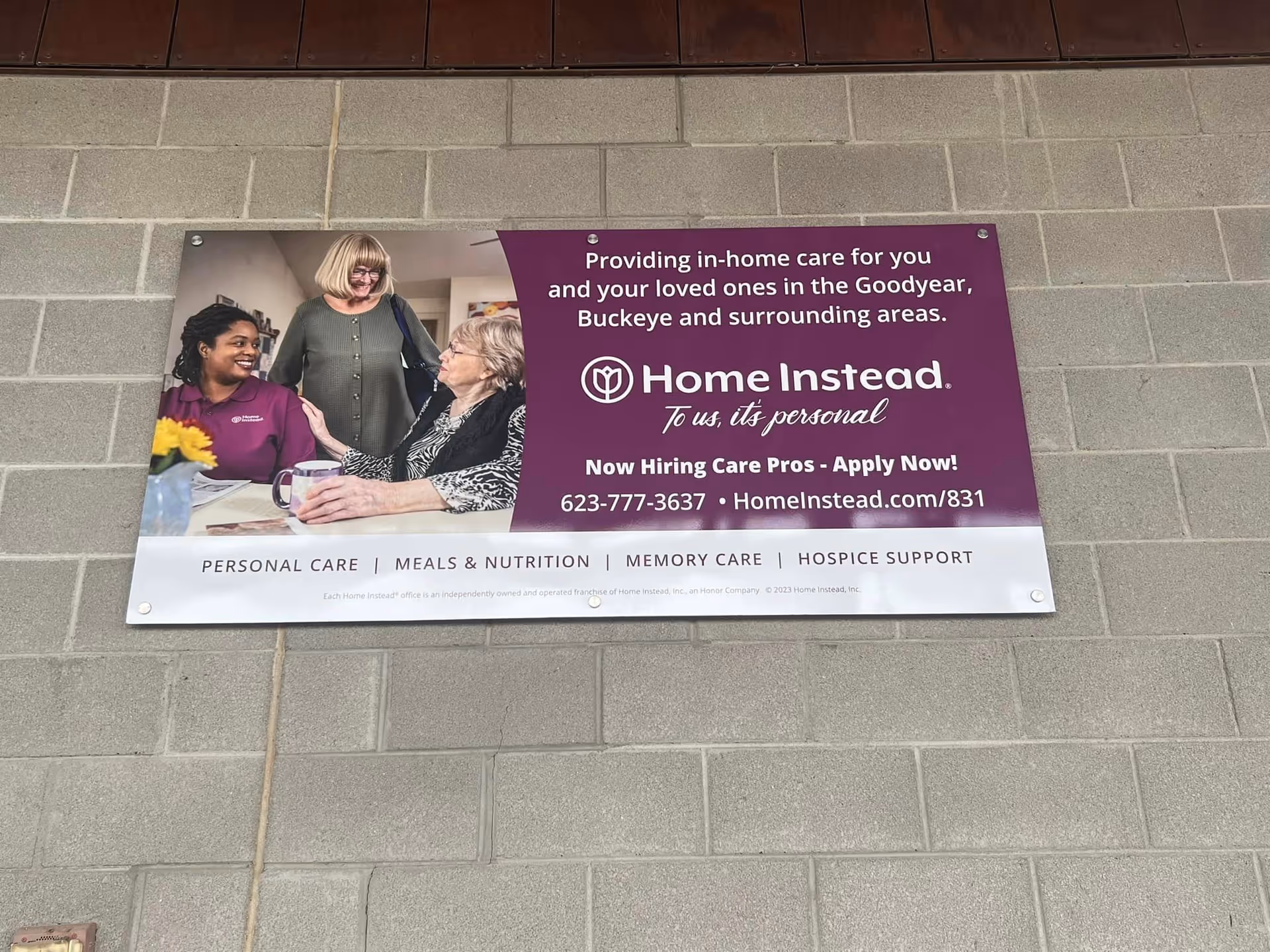 A Home Instead sign mounted on a gray brick exterior wall featuring people in the graphic.