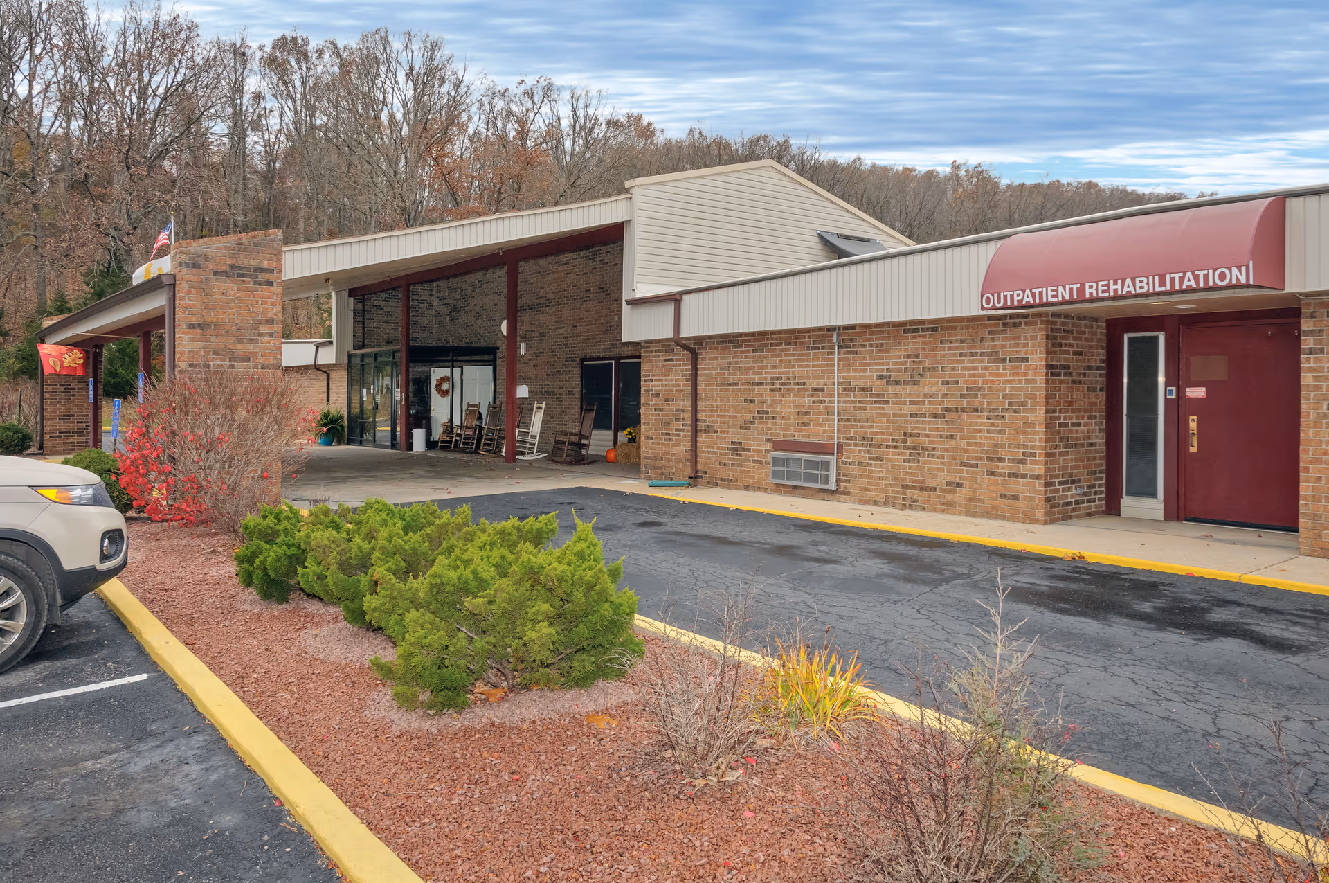 Exterior view of a brick building with a covered entrance and a red awning labeled 'OUTPATIENT REHABILITATION'. There are some shrubs and small trees in a landscaped area next to a parking lot with a vehicle partially visible. The sky is partly cloudy and there are leafless trees in the background.
