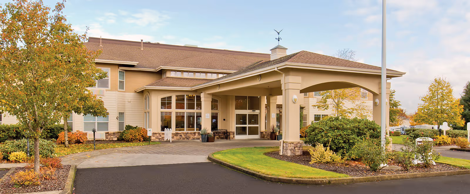 Exterior view of Brookdale Rose Valley Scappoose senior living facility showing the main entrance with a covered drop-off area, surrounded by landscaped greenery and trees with autumn foliage under a partly cloudy sky.