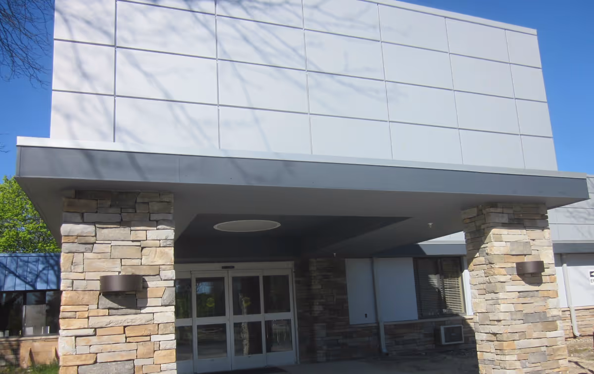 Entrance of a building with stone pillars supporting a gray overhang above automatic glass doors, under a clear blue sky.