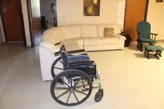 A wheelchair positioned in front of a beige sectional sofa in a room with light-colored flooring. To the right of the sofa is a green rocking chair. The room has light-colored walls with a framed picture hanging above the sofa and two doors visible in the background.