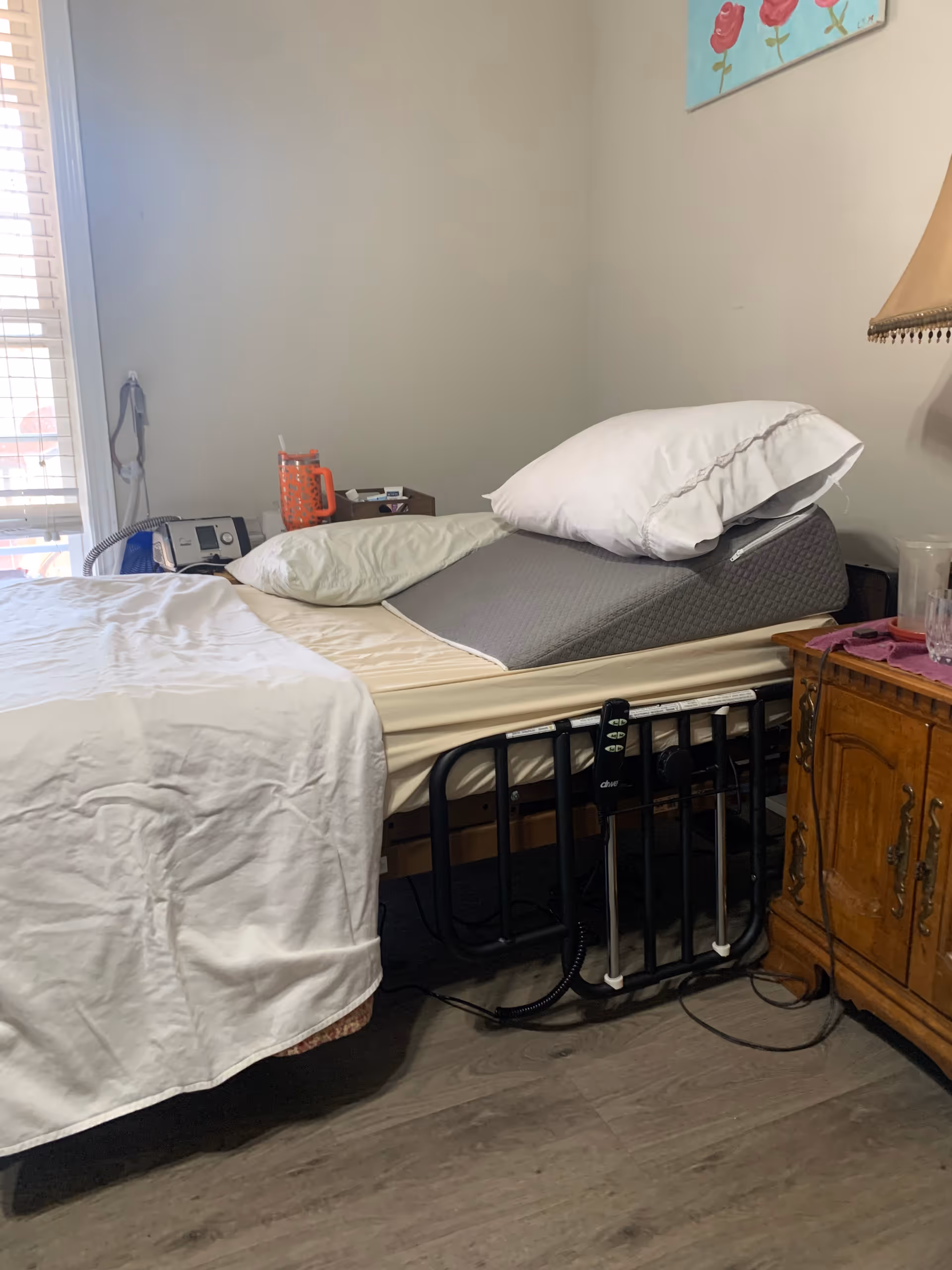 A single hospital-style bed in a small bedroom with a dresser, pillows, and window blinds.