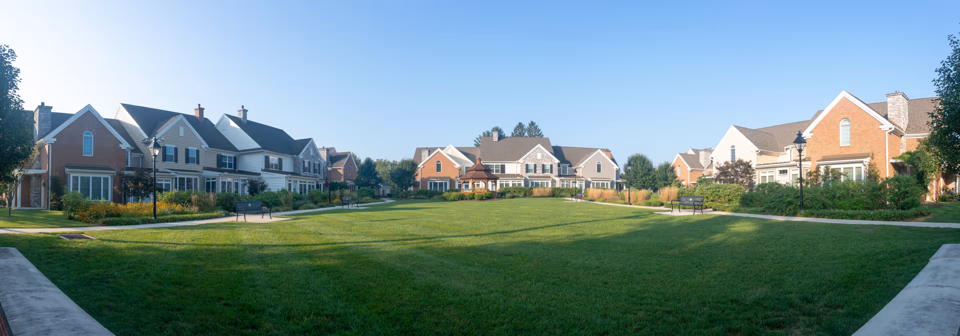 A panoramic view of a residential community with multiple two-story houses surrounding a large, well-maintained grassy courtyard. The houses have a mix of brick and siding exteriors, with several windows and chimneys visible. There are paved walkways, benches, and street lamps around the courtyard under a clear blue sky.