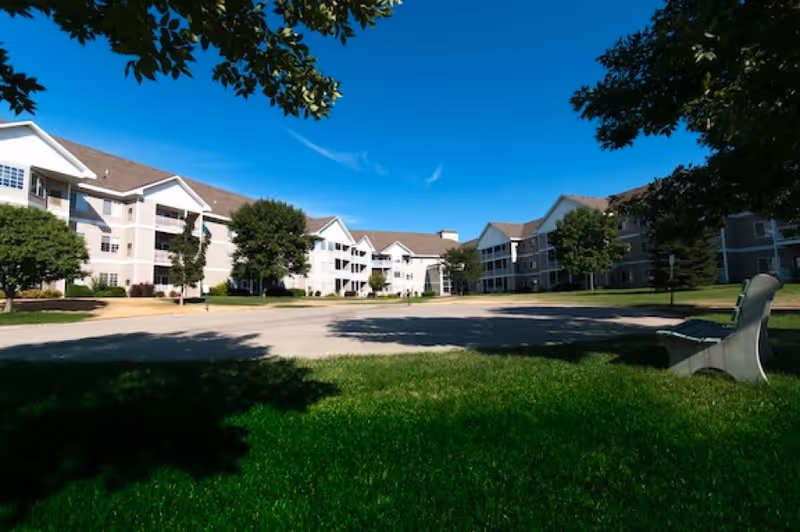 Exterior view of a senior living community with multiple three-story buildings surrounded by green lawns and trees under a clear blue sky. A bench is visible on the right side in the shaded grass area.