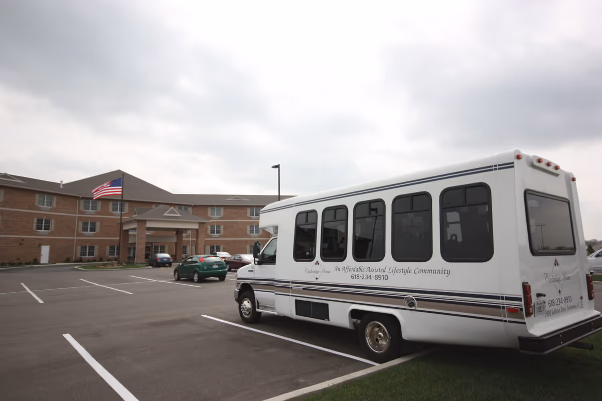 A white shuttle bus parked in a parking lot in front of a three-story brick building with an American flag flying on a flagpole. The bus has text on its side indicating it is from Cambridge House, an affordable assisted lifestyle community. Several cars are also parked in the lot under a cloudy sky.