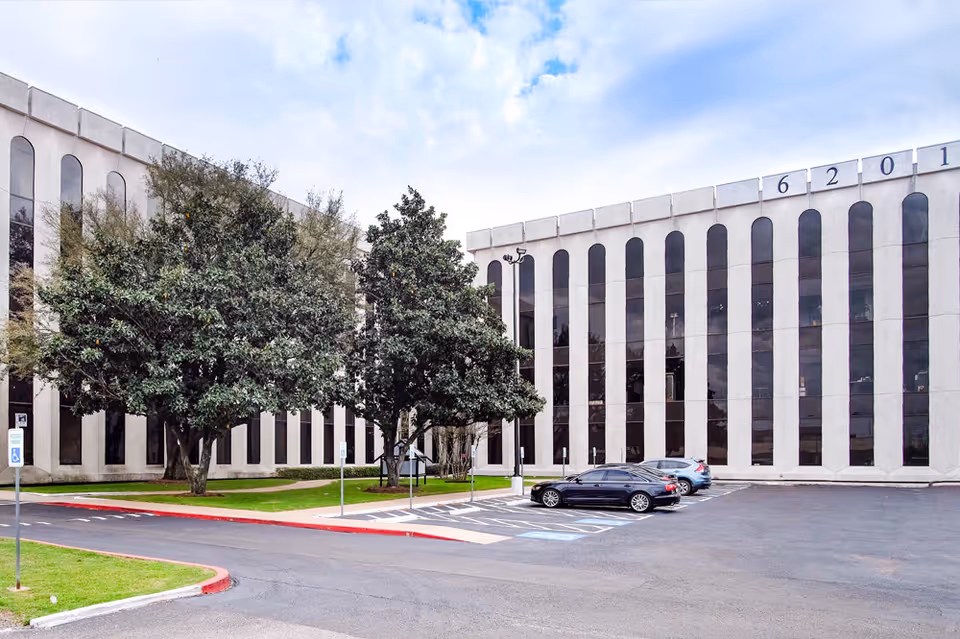 Large building exterior with tall vertical windows, a parking lot, and trees in front.