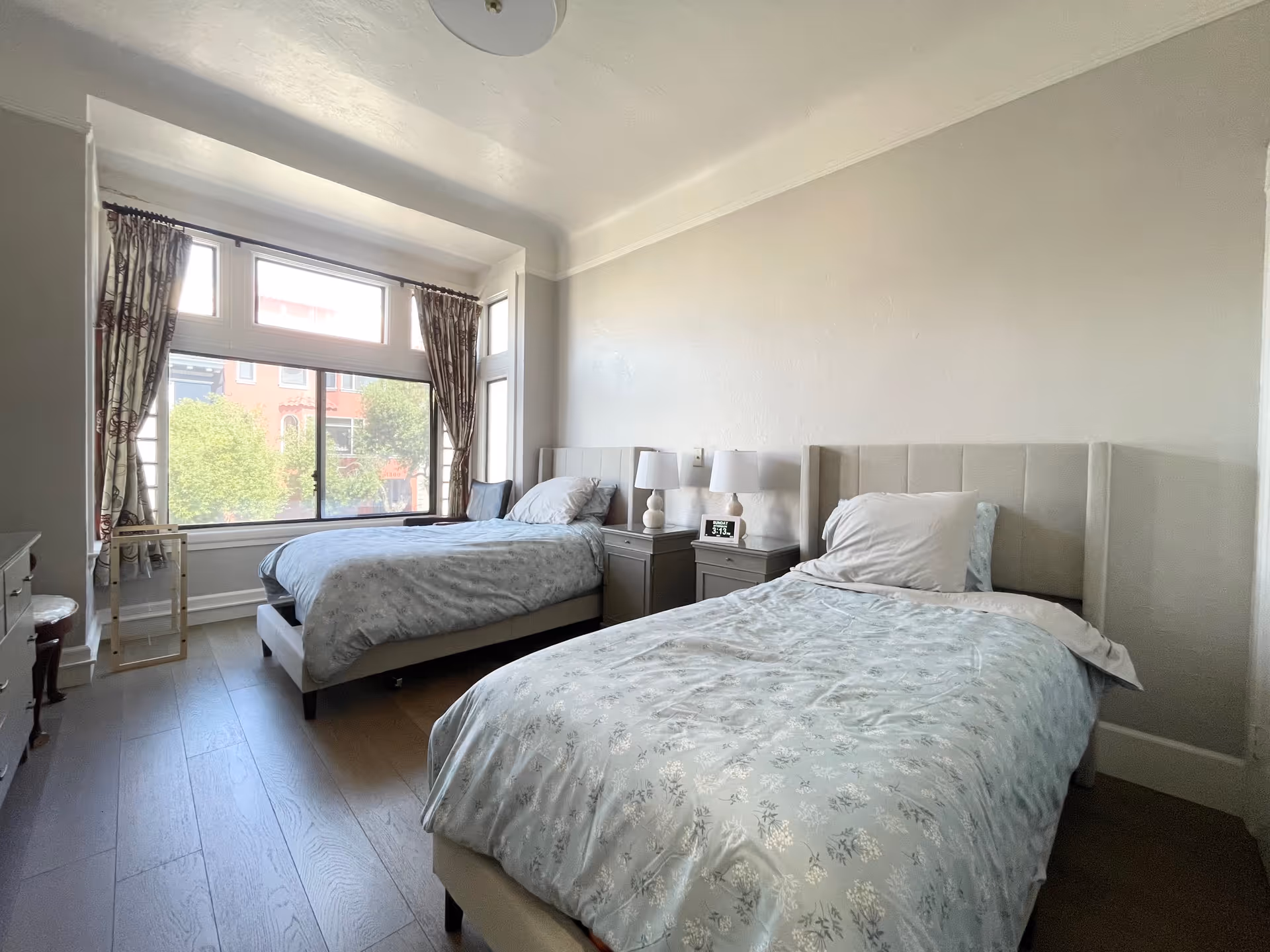 A bright bedroom with two single beds, each with light-colored bedding and pillows. Between the beds are two bedside tables with matching lamps and a digital clock. A large window with patterned curtains lets in natural light, and the room has light-colored walls and wooden flooring.
