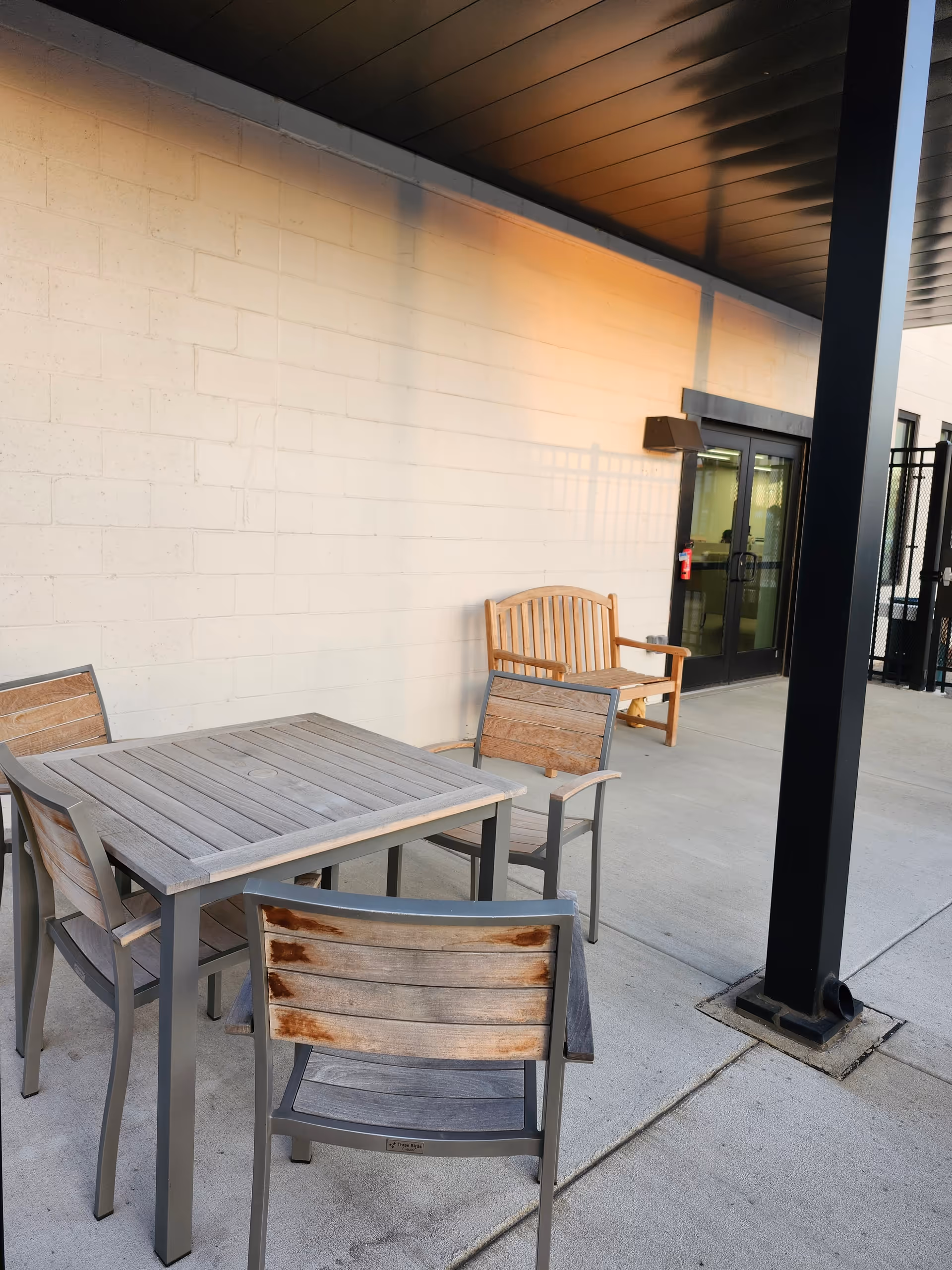 Outdoor patio with wooden table, chairs, and a bench outside the building.