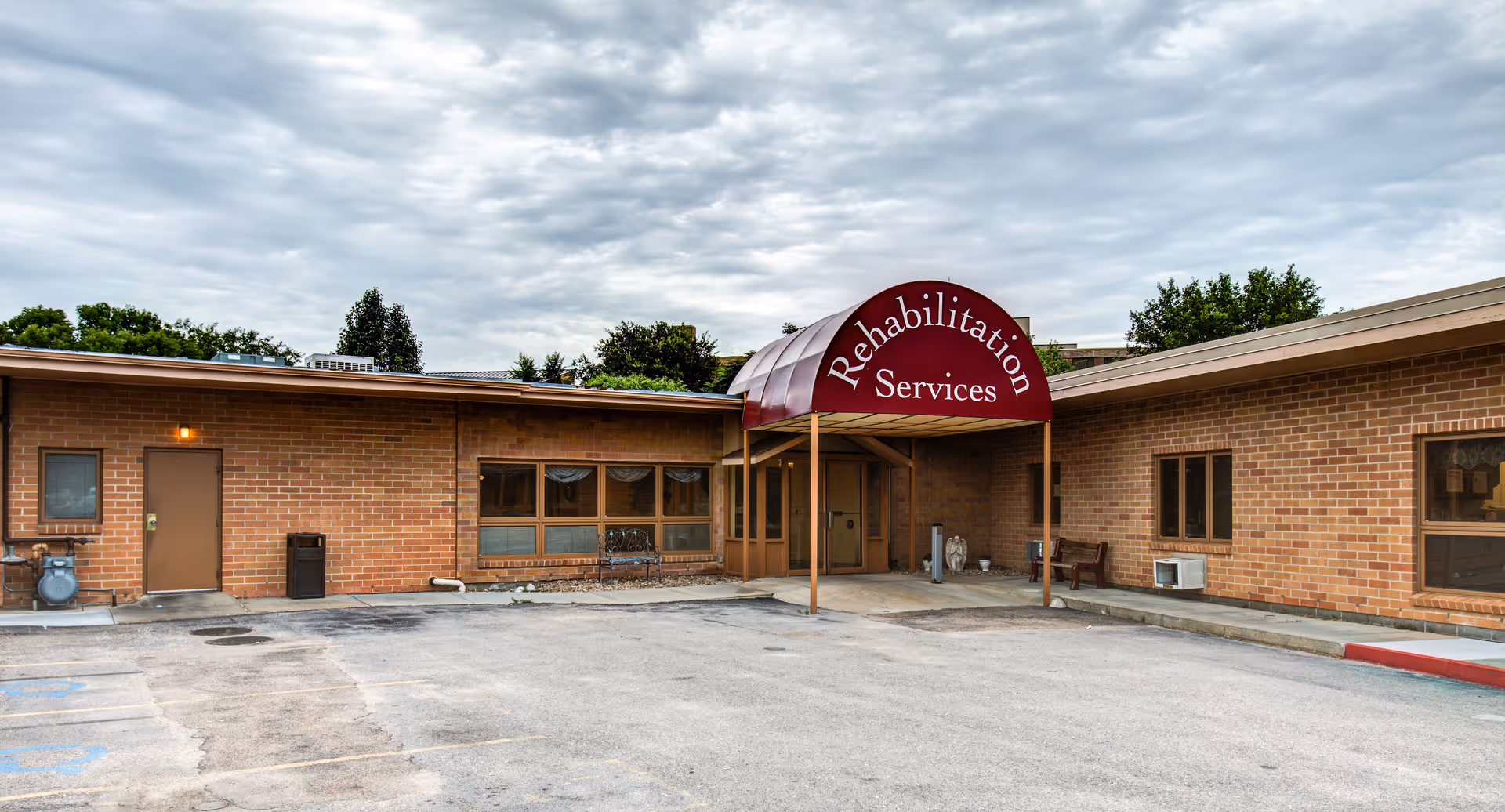 Single-story brick building entrance with a maroon awning reading "Rehabilitation Services," an empty parking area, and a cloudy sky.