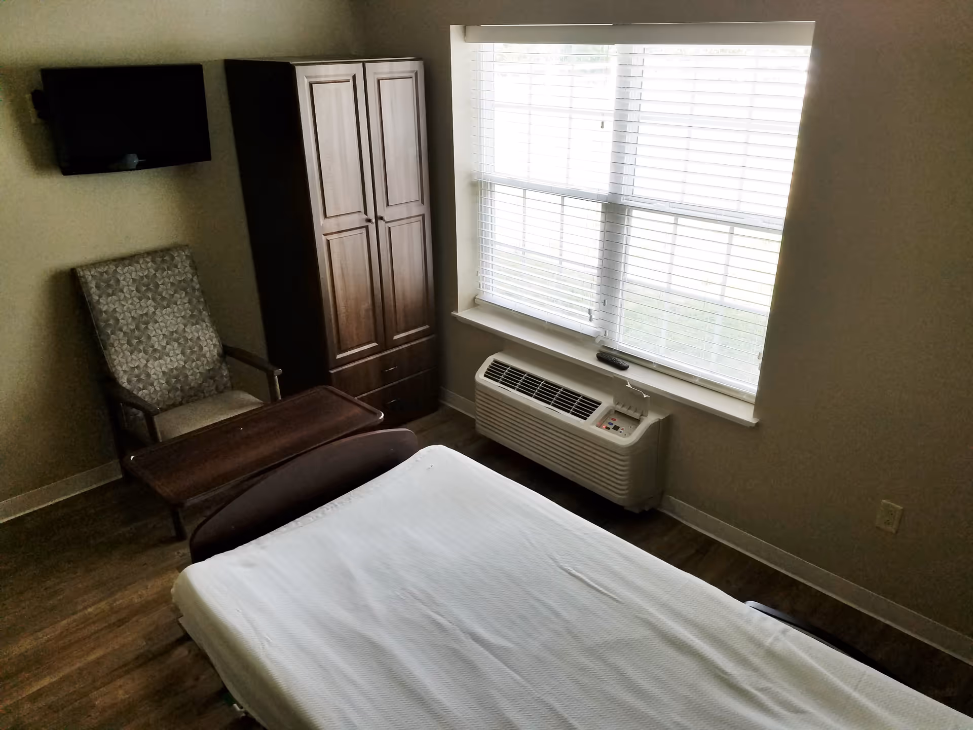 A bedroom with a bed, chair, wardrobe, large window with blinds, and a window air conditioner.