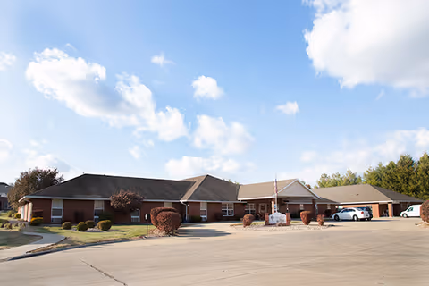 Single-story brick building with a dark roof under a blue sky with scattered clouds. The building has several windows and a covered entrance with an American flag. There are trimmed bushes and a paved driveway with parked cars in front.