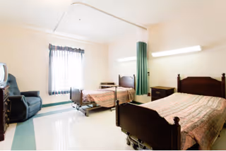 Two hospital-style beds in a bright patient room with window and chair.