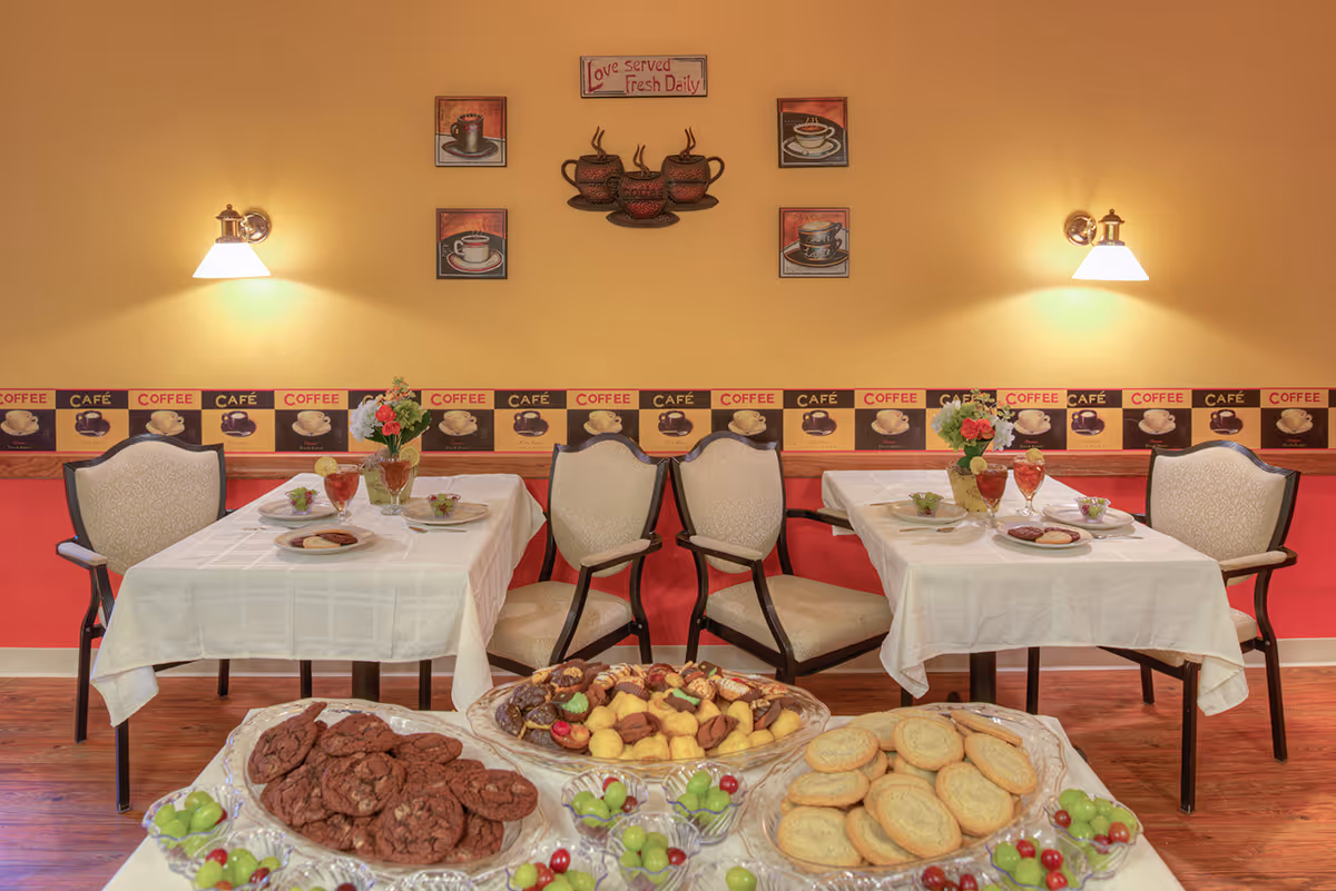 Two small dining tables with white tablecloths and place settings in a cozy dining room with coffee-themed wall decor and platters of cookies and desserts in the foreground.
