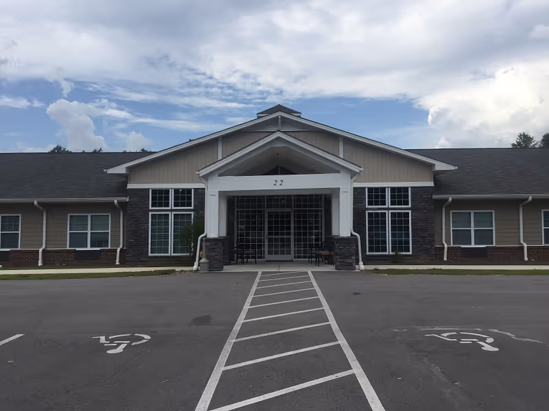 Front exterior view of a single-story building with a covered entrance supported by white columns. The building has large windows and a combination of stone and siding on the facade. There are two handicap parking spaces in front of the entrance, and the sky is partly cloudy.