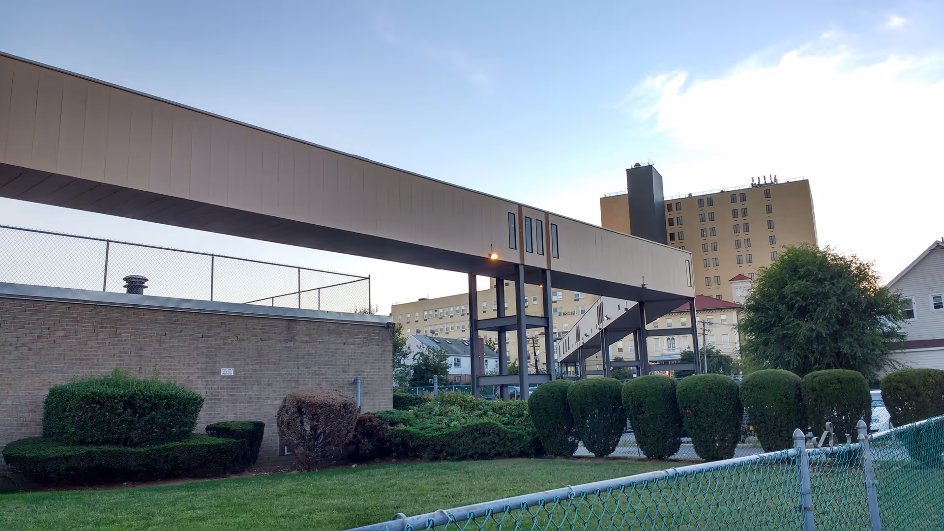 Exterior view of an elevated walkway connecting buildings with neatly trimmed hedges and a grassy lawn.