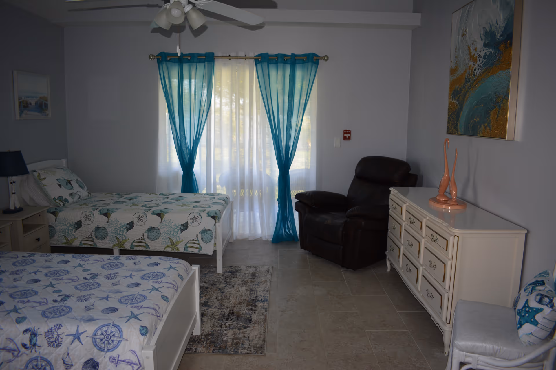 Shared bedroom with two beds, turquoise curtains, dresser, and a recliner.
