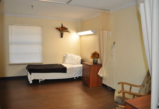 Nursing home bedroom with single bed, dresser, lamp, flowers, cross on wall, privacy curtains.