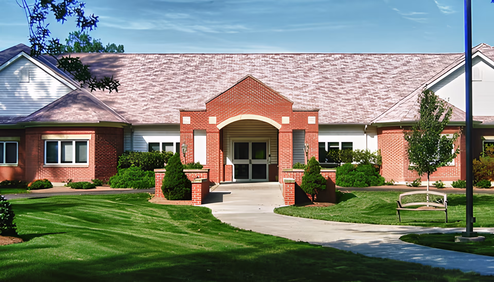Front entrance of a single-story brick retirement community building with a paved walkway, manicured lawn, and landscaping.