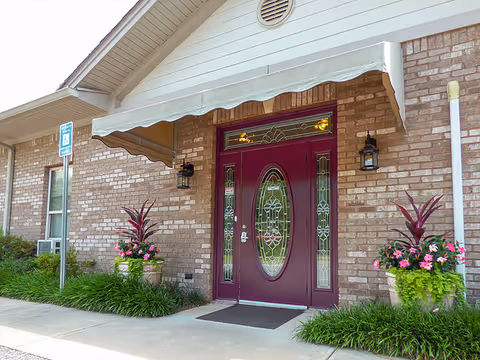 Brick building entrance with a maroon oval-glass front door, awning, wall lanterns, and potted flowers.