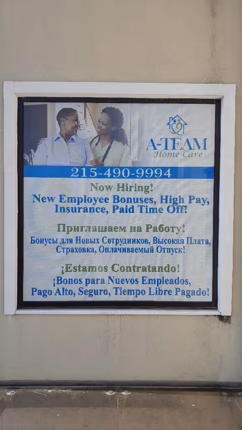 Framed A-TEAM Home Care recruitment sign with two people on an exterior building wall.