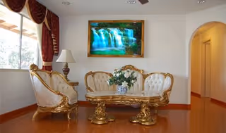 Ornate gold furniture surrounds a glass coffee table in a bright living room with waterfall art