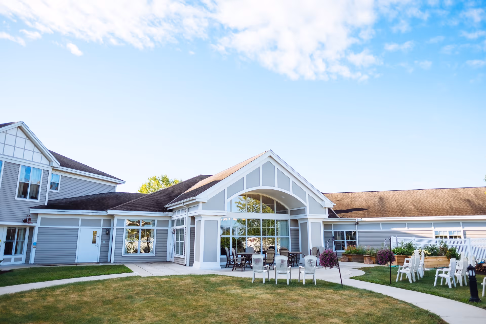 Exterior view of a senior living facility building with gray siding and white trim. The building has a peaked roof and large windows. In front of the building is a grassy area with a curved concrete walkway and several white chairs arranged around tables and fire pits. The sky is clear with some scattered clouds.