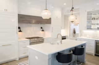 A modern white kitchen with an island, pendant lights, and stainless steel appliances.
