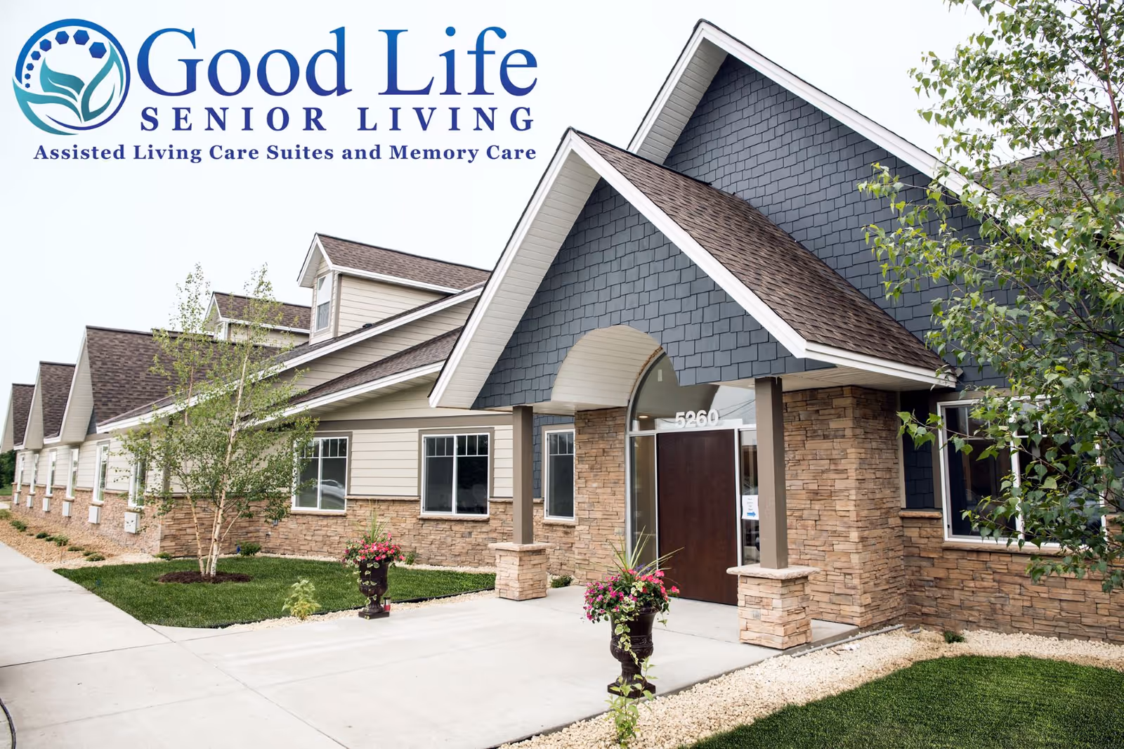 Front entrance of Good Life Senior Living facility with stone facade, pitched roof, landscaped walkway, and the Good Life Senior Living logo at the top left.