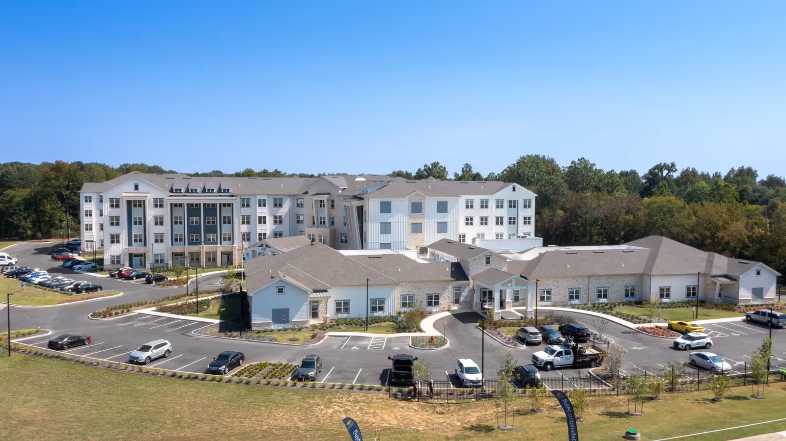 Exterior view of The Summit of Germantown senior living facility showing a large multi-story building with white and gray siding, multiple windows, and a parking lot with several cars parked. The building is surrounded by landscaped greenery and trees under a clear blue sky.