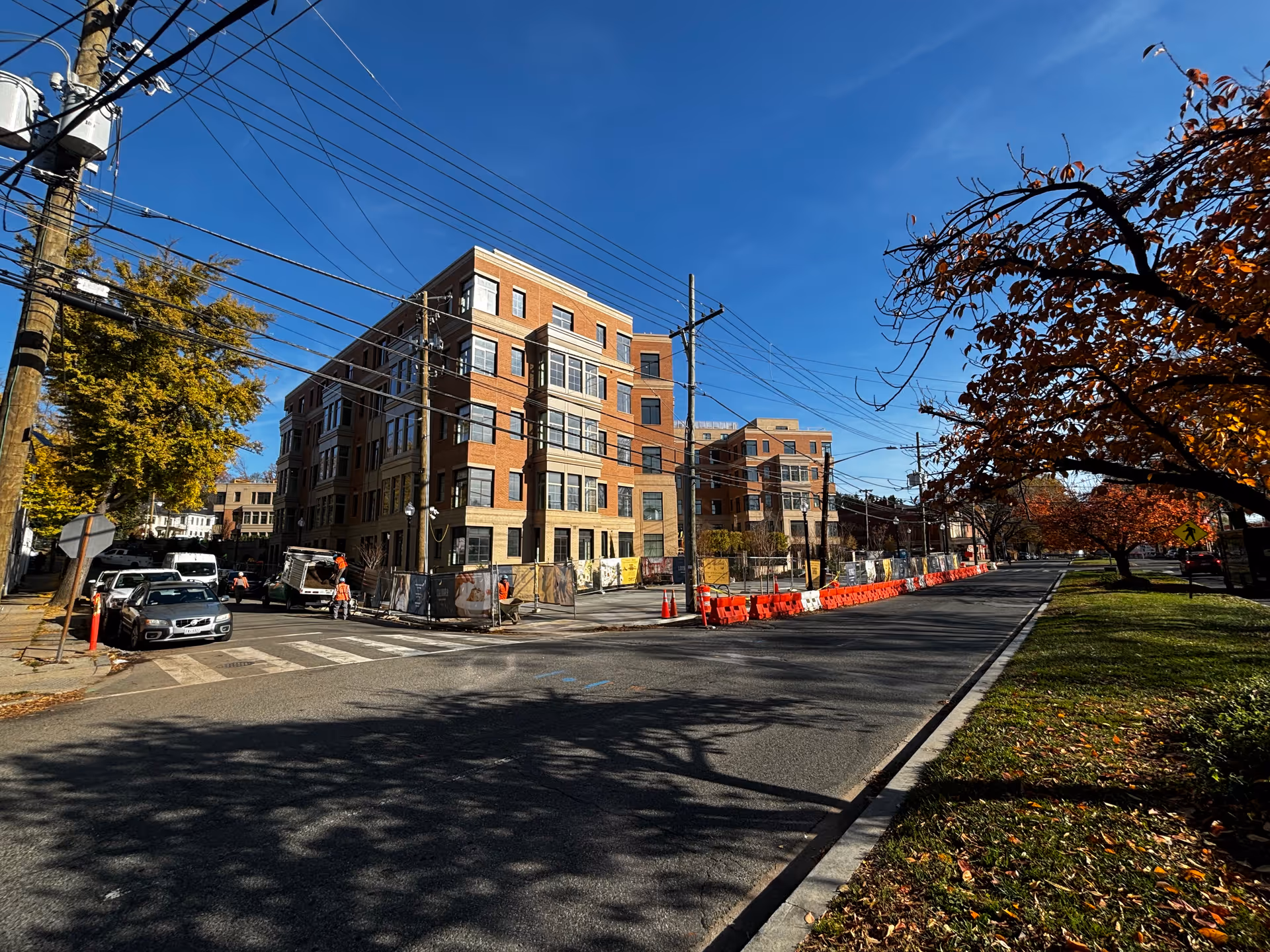 Multi-story brick senior living building along a city street with construction barriers, orange cones, parked cars, and trees with autumn leaves.