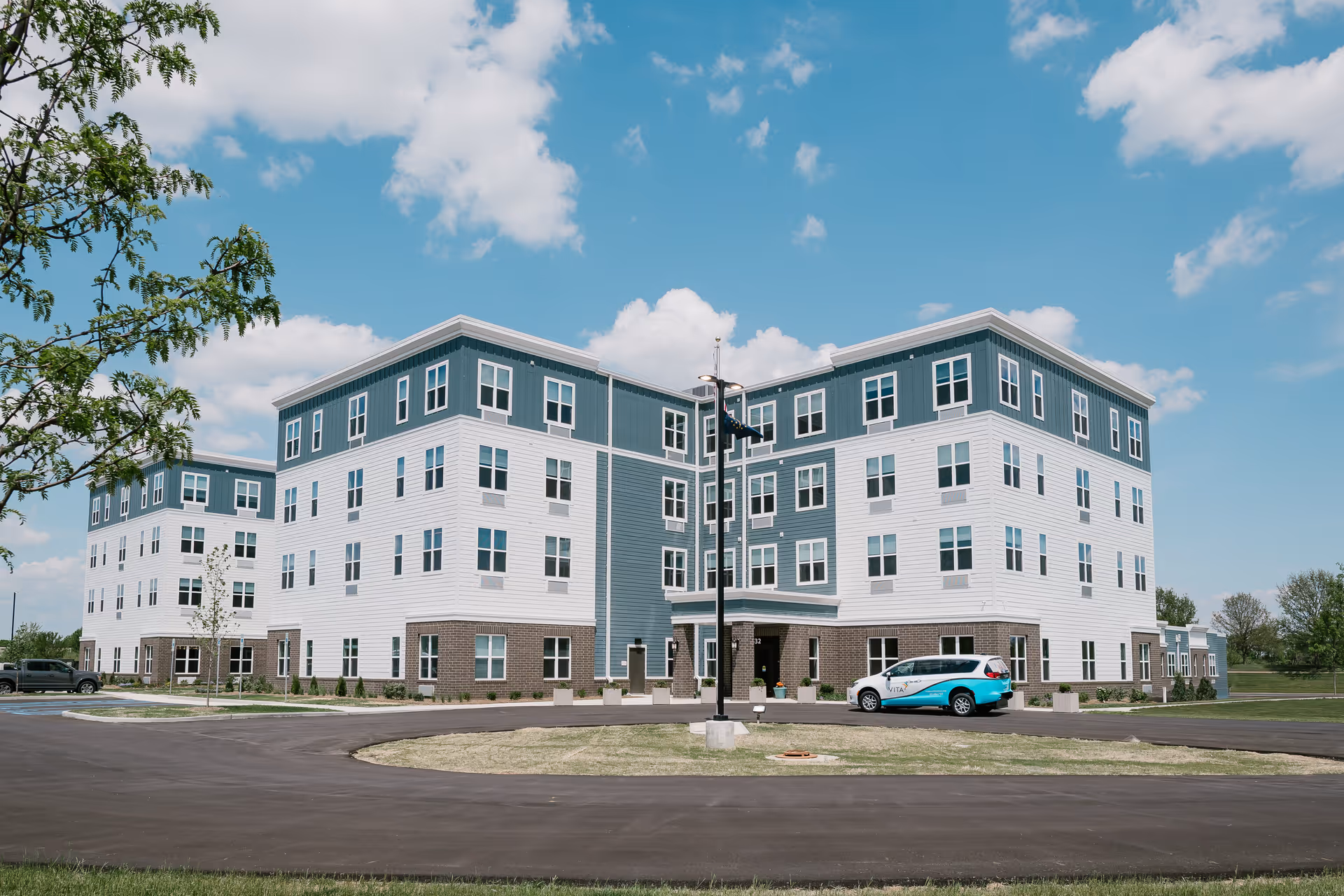 Exterior view of a modern four-story senior living facility building with a combination of white and blue siding and brick accents on the lower level. A circular driveway is in front with a parked vehicle branded with 'Vita'. The sky is partly cloudy and there are some trees and grass around the building.