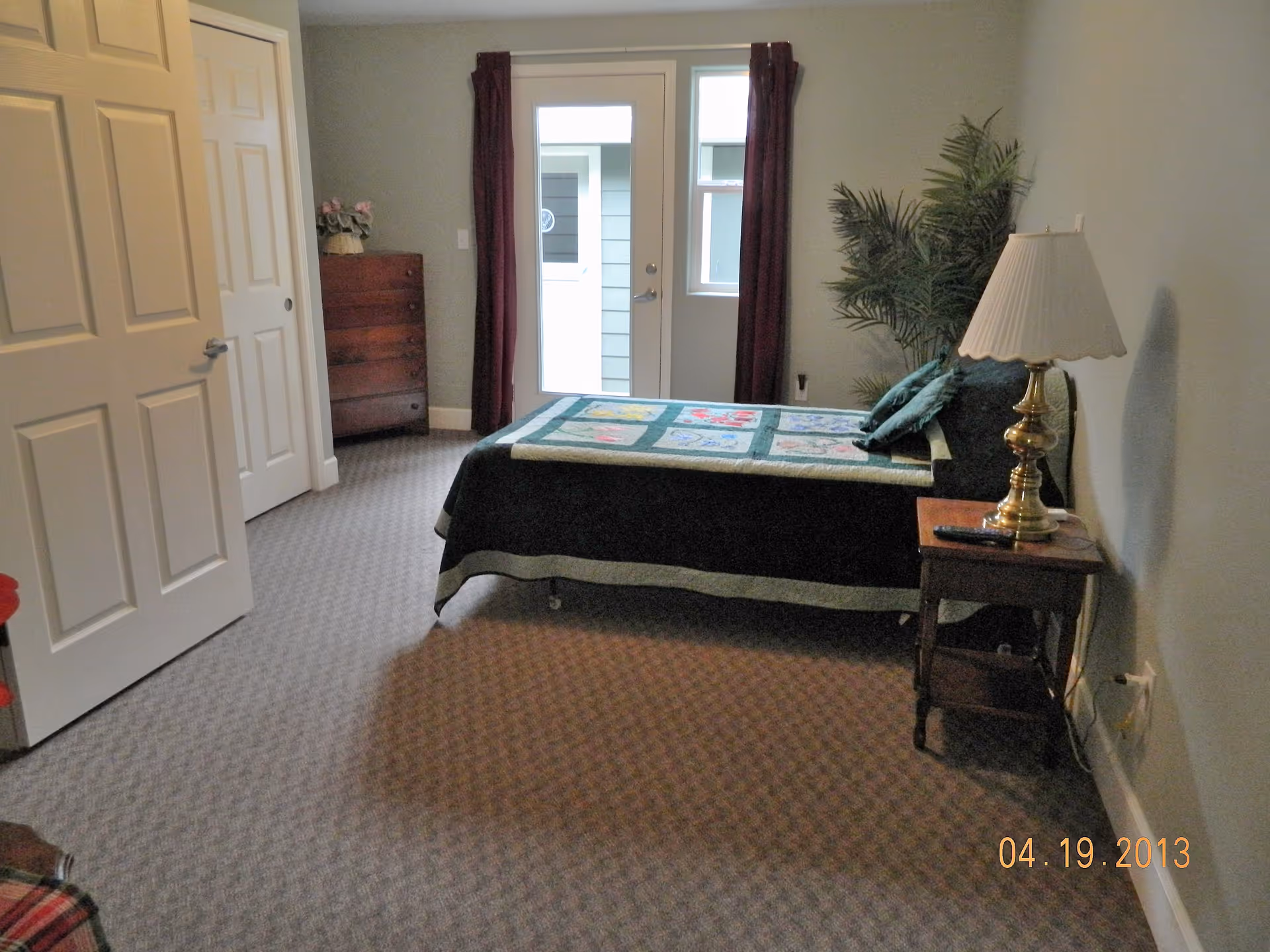 A bedroom with a single bed covered with a colorful quilt, a wooden nightstand with a brass lamp, a tall wooden dresser with a flower vase, a potted plant in the corner, and a door with a window leading outside. The room has carpeted flooring and light-colored walls.