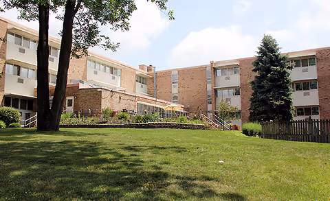 Exterior view of a multi-story brick retirement community building with large windows, surrounded by green grass, trees, and a clear blue sky with some clouds.