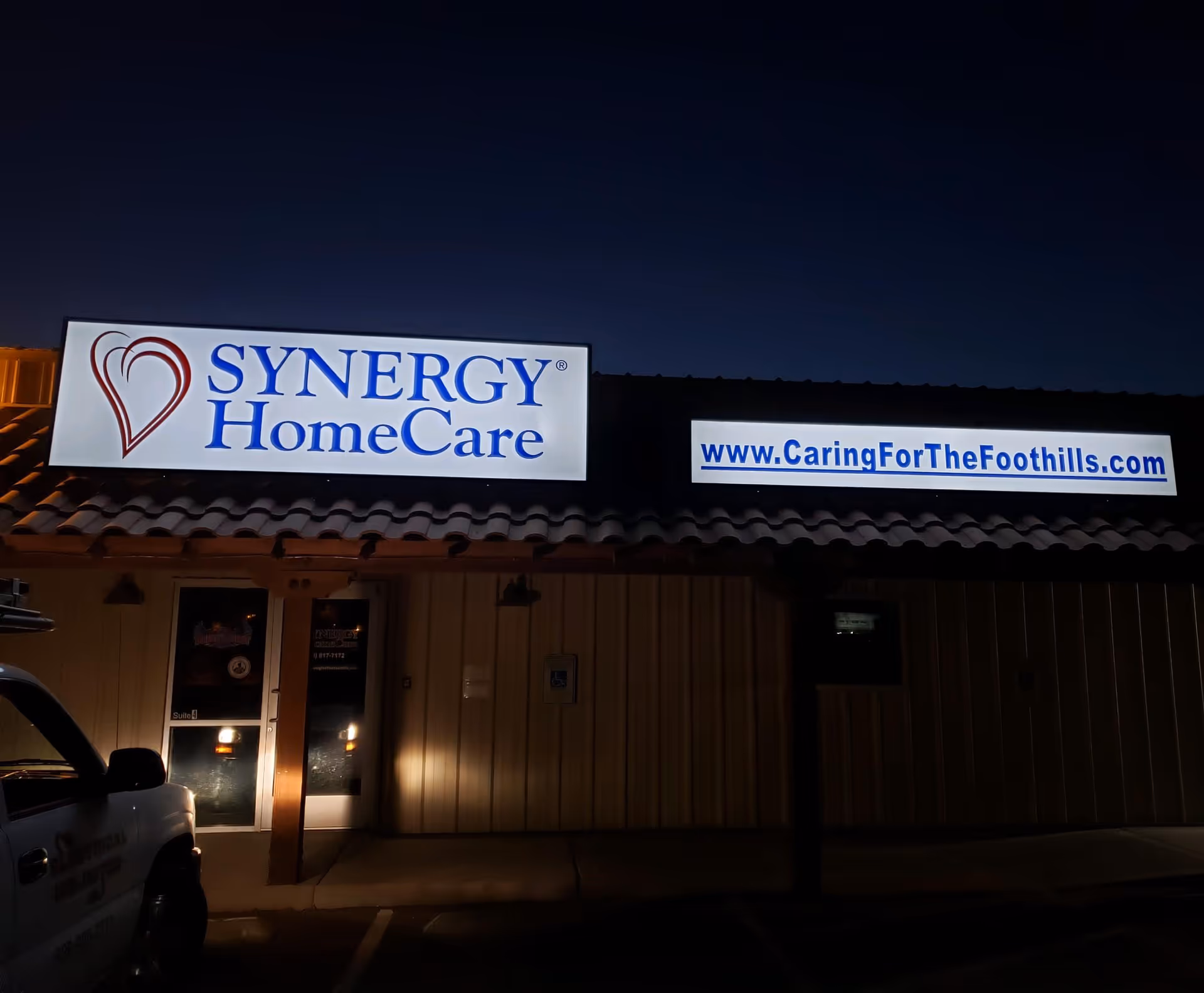 Night exterior of a building with illuminated SYNERGY HomeCare and CaringForTheFoothills signs.