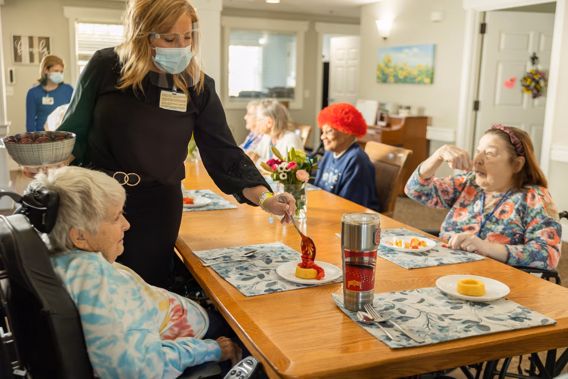 A masked staff member wearing a face shield serves dessert to elderly residents seated around a dining table in a senior living dining room.