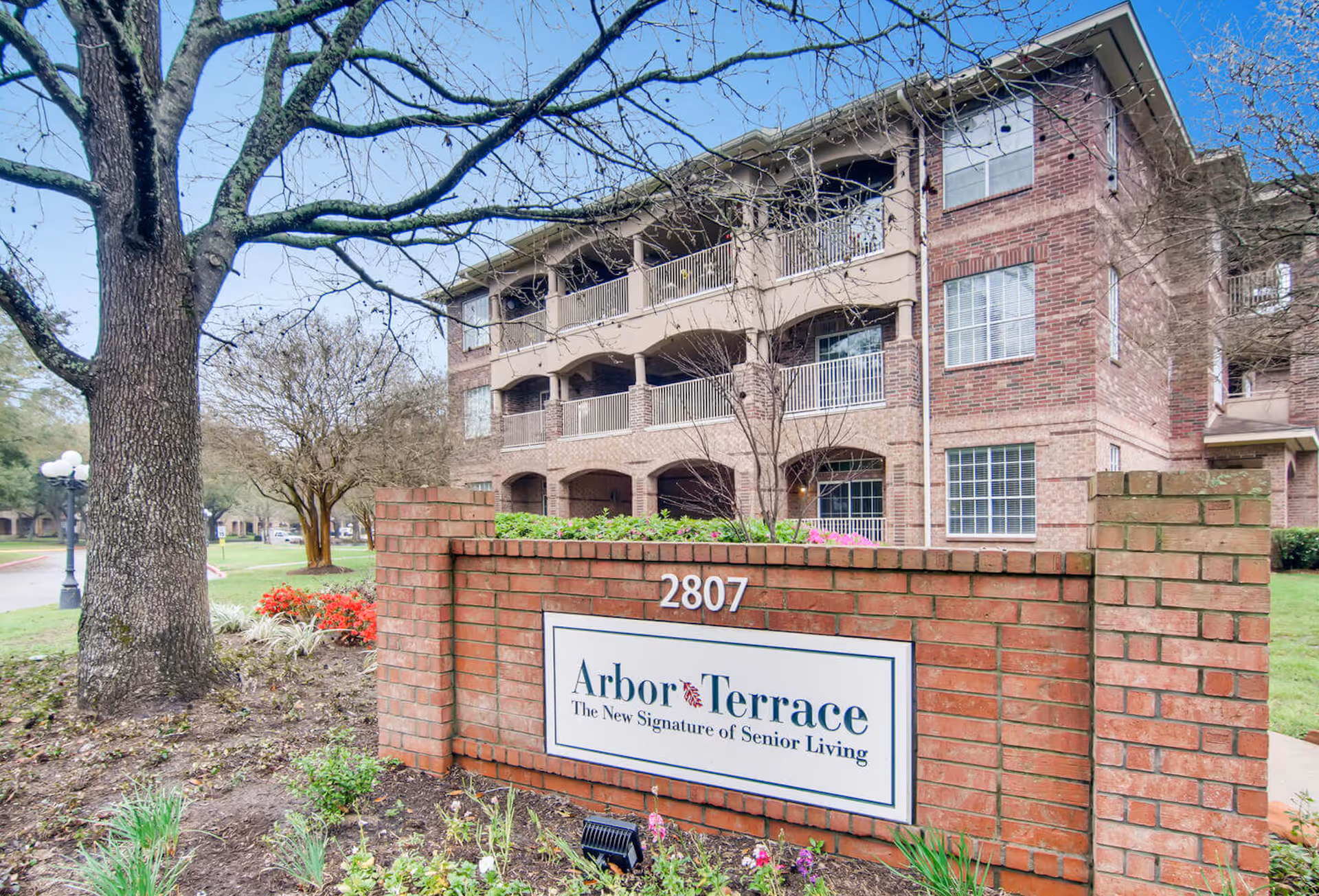 Three-story brick senior living building with covered balconies and a brick entrance sign reading 'Arbor Terrace' and address 2807.