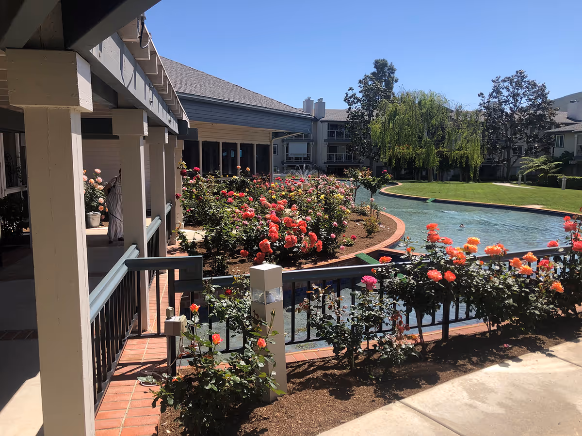 Courtyard with rose beds and a winding pond surrounded by covered walkways and senior living buildings under a clear sky.