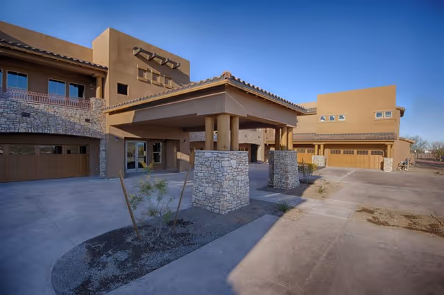 Exterior view of a modern senior living facility building with stone and stucco walls, a covered entrance supported by stone pillars, multiple garage doors, and a clear blue sky.