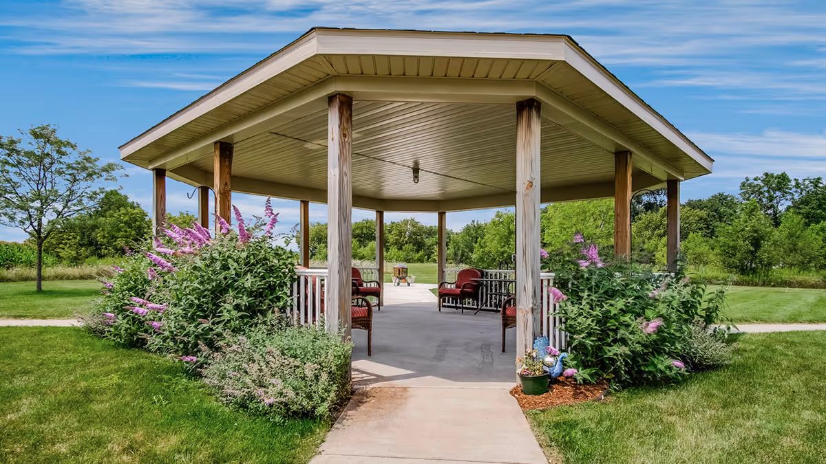 Wooden octagonal gazebo with chairs surrounded by flowering shrubs and a grassy lawn under a blue sky.