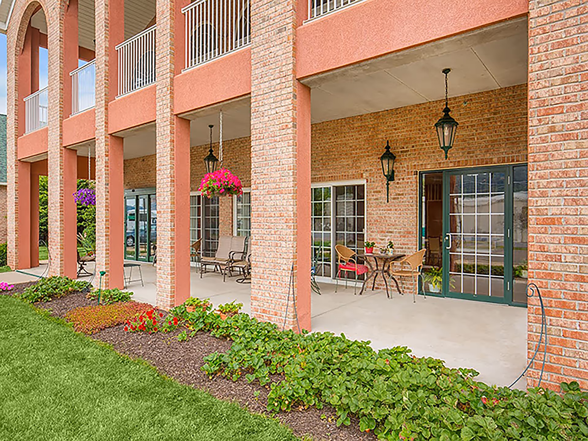 Outdoor covered patio area of a brick building with hanging flower baskets, outdoor seating including chairs and tables, and green plants and grass in the foreground.