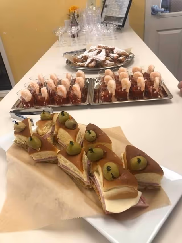 A countertop with trays of food including small sandwiches topped with green olives, shrimp cocktail in individual glasses, and a plate of powdered sugar-covered pastries. In the background, there are empty wine glasses and a menu or sign on a stand.