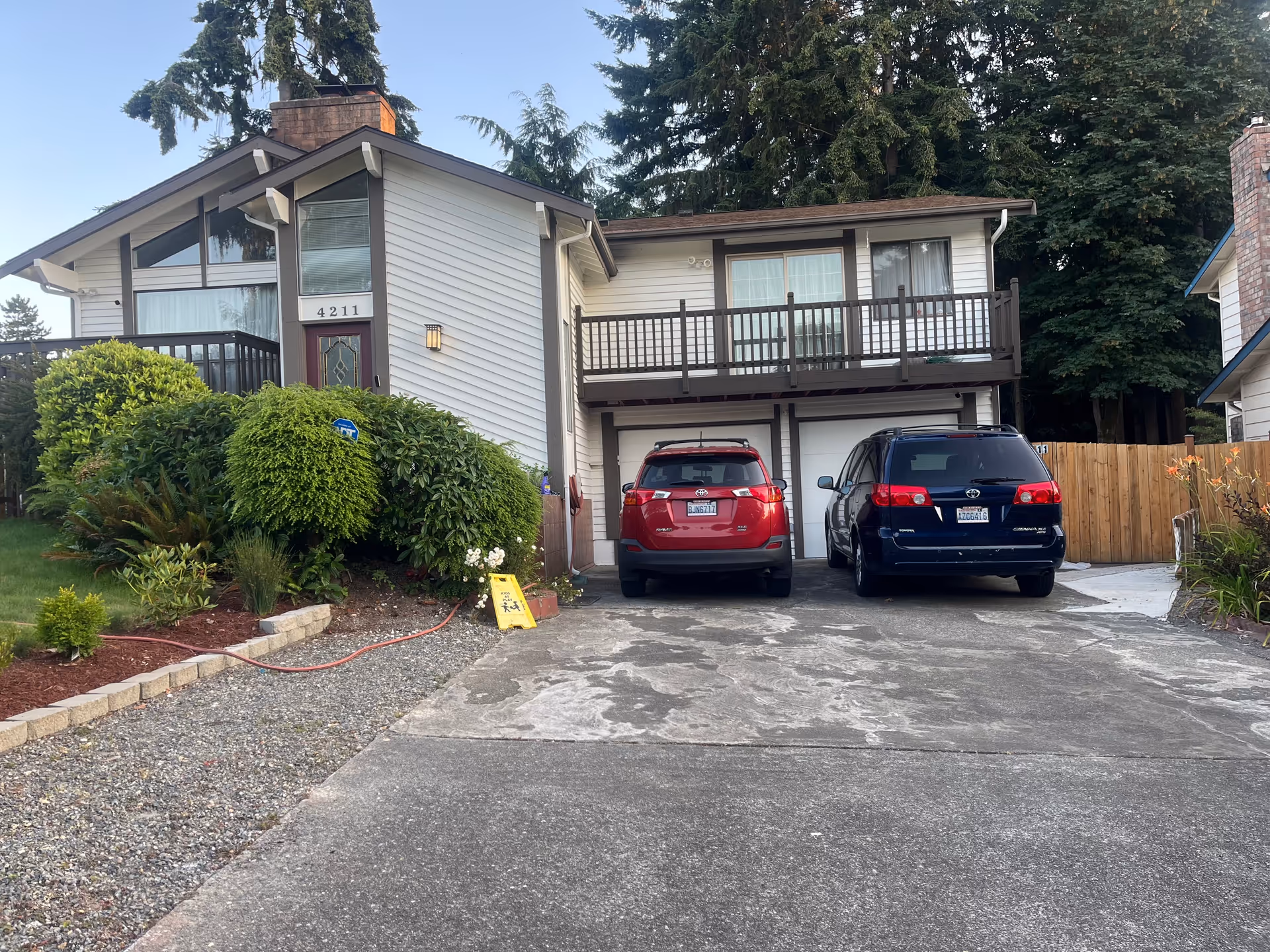 Two-story house-style senior living facility with a driveway, two parked SUVs, and shrubs.