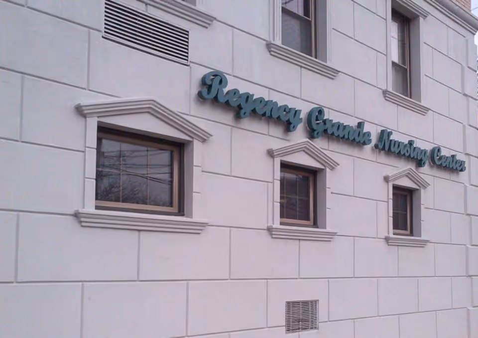 Exterior facade of a light-colored building with three small windows and green script signage reading 'Regency Grande Nursing Center'.