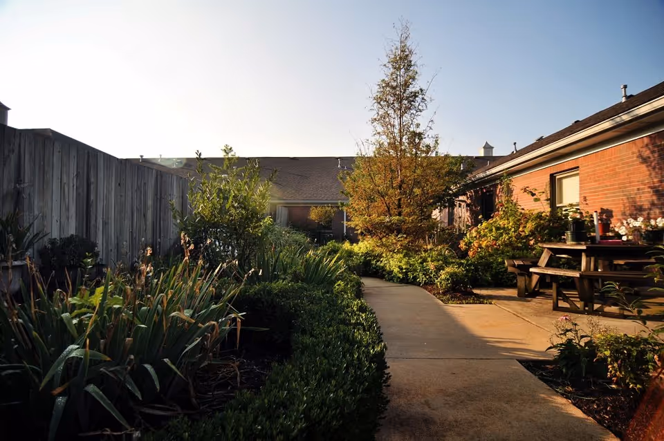 Sunlit courtyard path between brick buildings with garden beds, a tree, and picnic benches.
