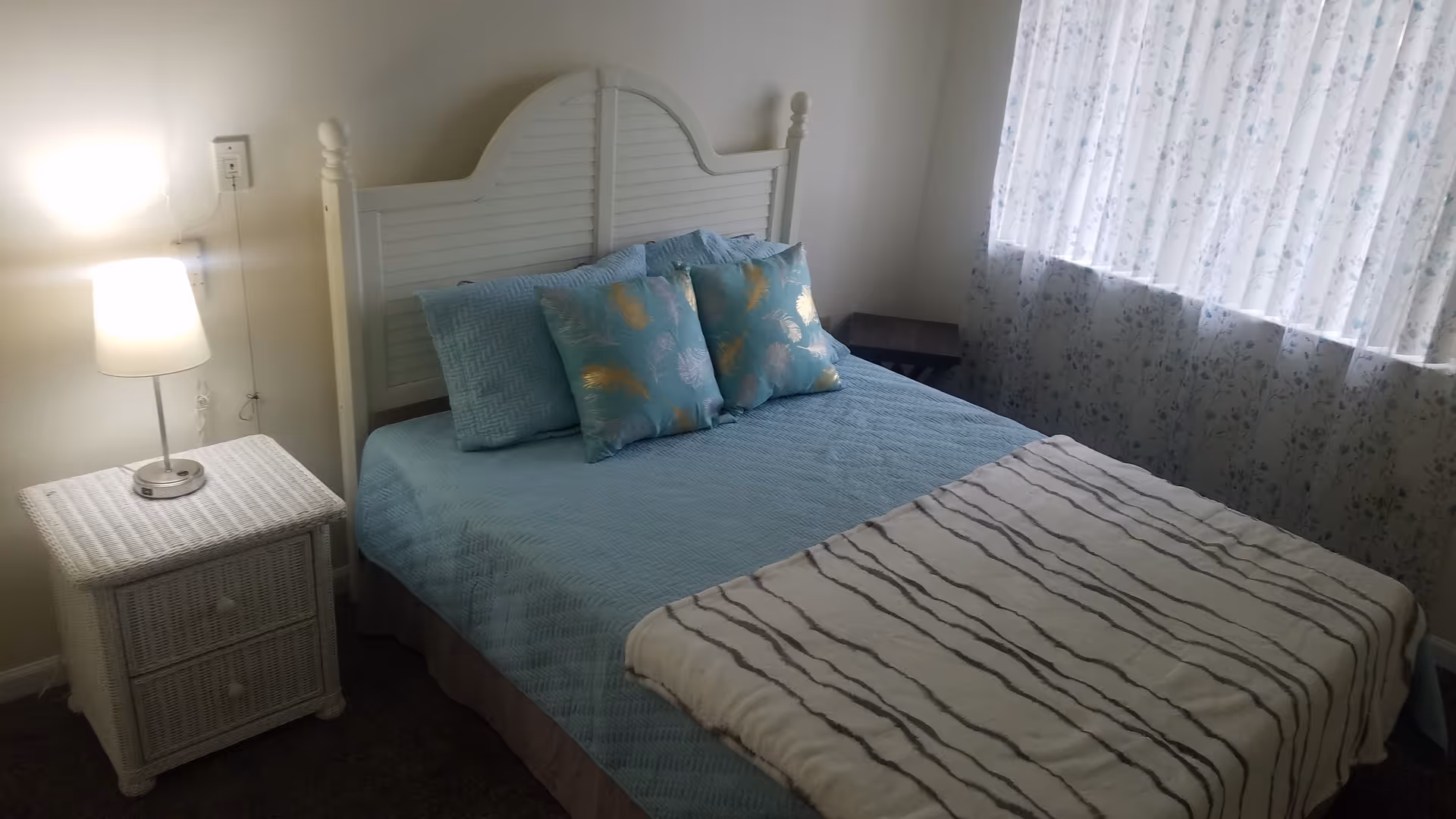 White headboard bed with blue bedding and wicker nightstand in a sunlit bedroom.