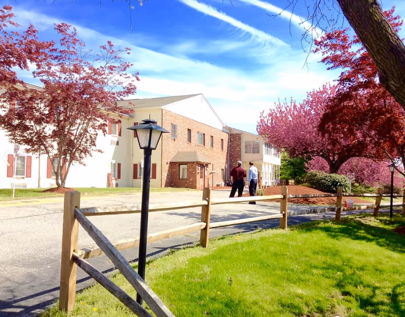 Exterior view of Allegria at The Oaks showing brick building, blossoming trees, fence, and two people.