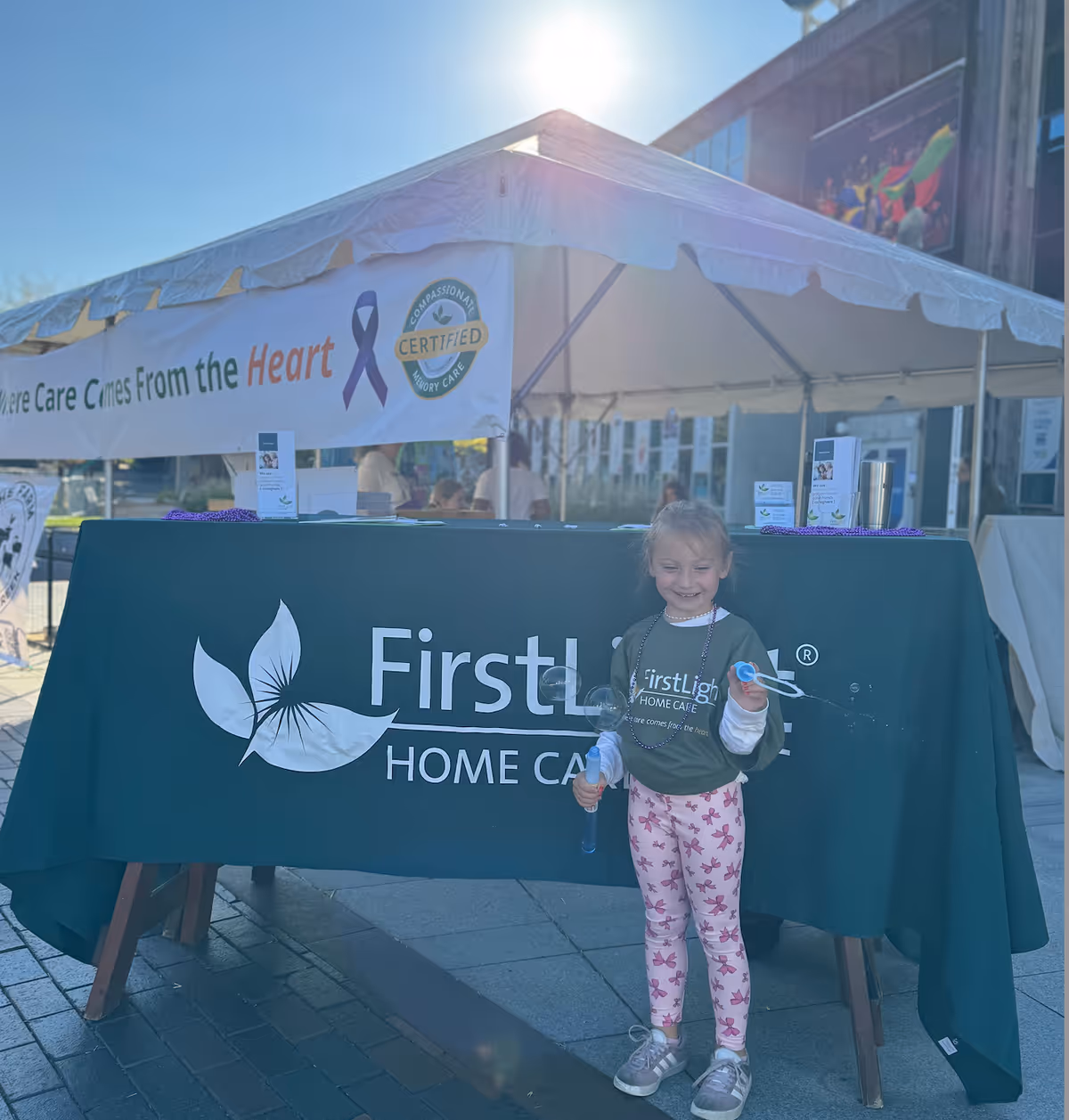 Outdoor FirstLight Home Care booth with a tent, banner, and a smiling child.