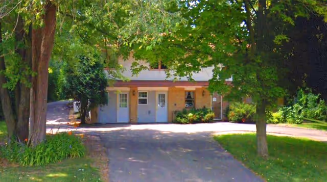Front view of a beige senior living building with multiple doors, trees, and a driveway.