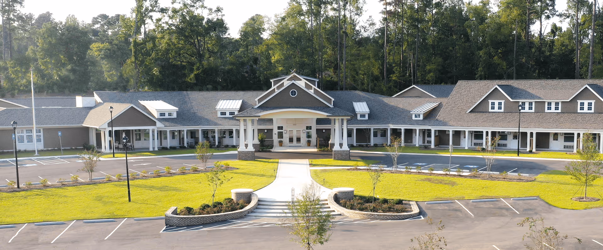 Front exterior view of a single-story senior living facility building with a covered entrance, surrounded by a well-maintained lawn, small trees, and a parking lot with marked spaces. Tall trees form a wooded backdrop behind the building.